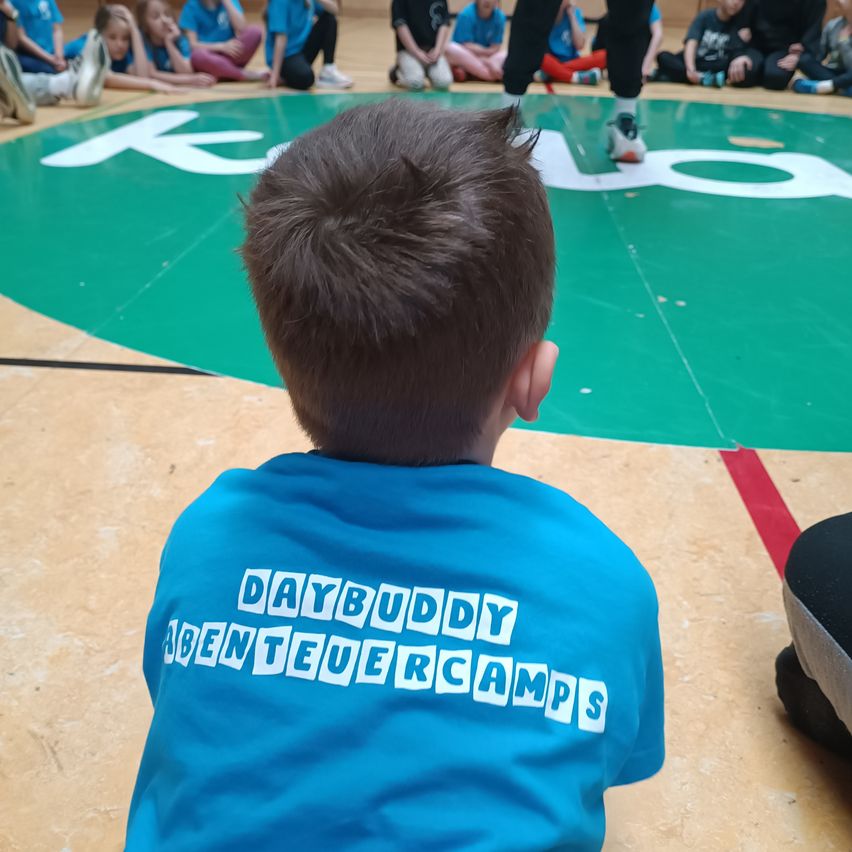 A young boy wearing a blue shirt with 'DAYBUDDY ABENTEUERCAMPS' printed on it is watching a wrestling match.