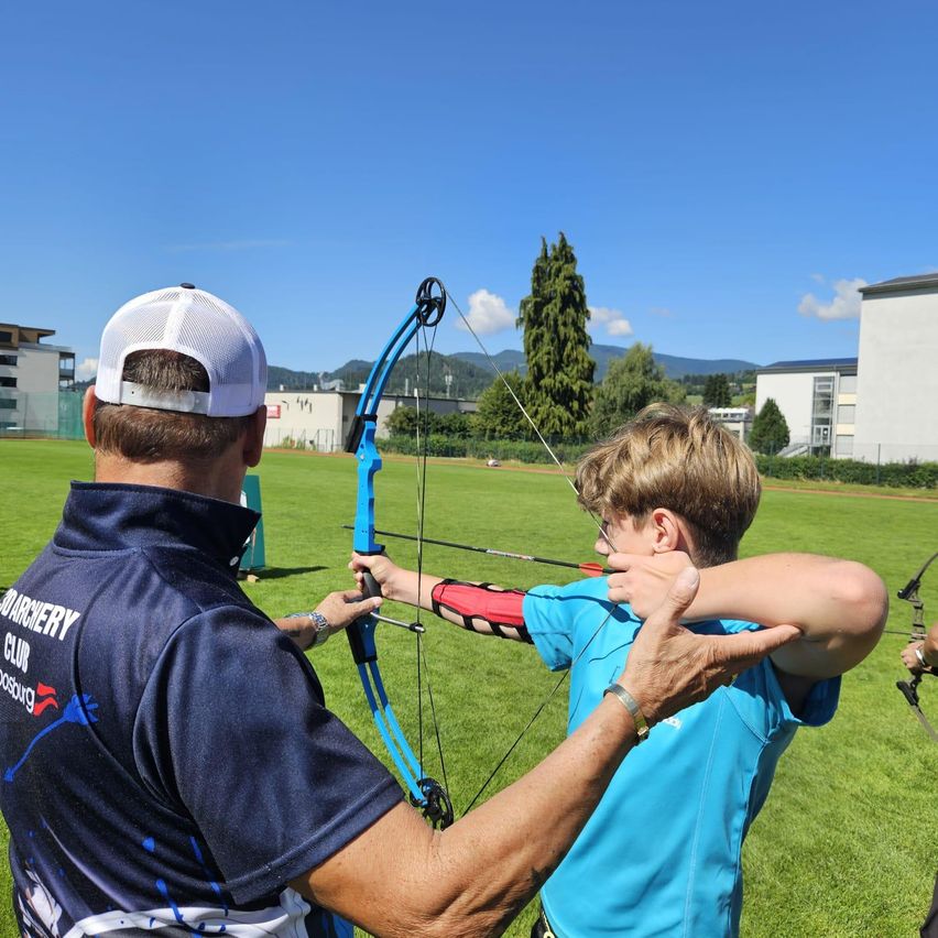 An adult man is teaching a young boy how to shoot a bow and arrow on a grassy field.