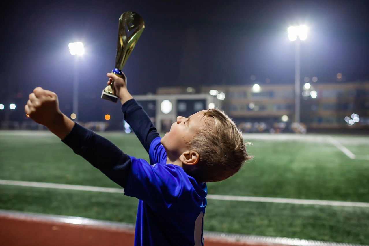 Ein junger Junge in einem blauen Shirt hält einen goldenen Pokal in die Höhe und schaut bei Nacht auf einem Sportplatz in den Himmel.