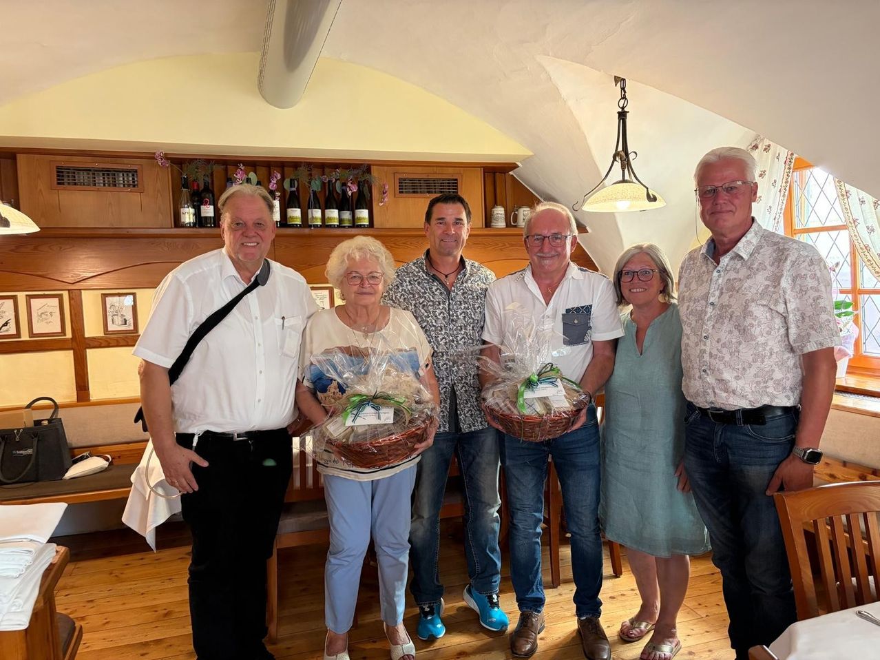 A group of six people standing together in a room, two of them holding baskets with gifts inside. They are all smiling and seem to be posing for a photo. Behind them is a wooden shelf with bottles and cups.