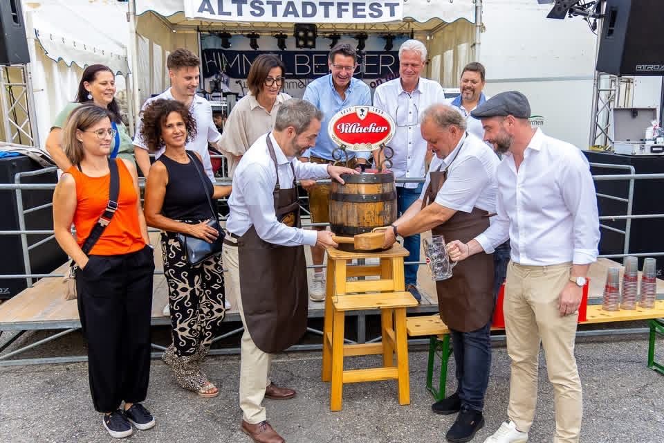 At the Altstadtfest, several men in aprons tap a barrel on a wooden stool while others watch. A banner for the event is visible in the background.