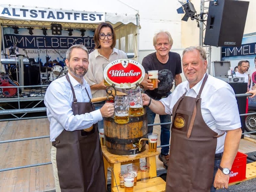 Four people are standing in front of a beer barrel with glasses. A woman and a man are wearing brown aprons, while the other two men are in casual attire. Behind them, a stage is set up with musical instruments and a banner.