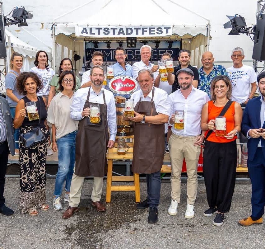 A group of people wearing aprons and holding beer mugs are posing for a photo at an event. A banner with the words Altstadtfest is hanging behind them.
