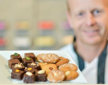 Ein Mann in einem weißen Hemd und einer schwarzen Krawatte hält einen weißen Teller mit einer Vielzahl von Gebäck, darunter Donuts, Schokoladen und Kekse.