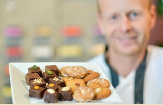 Ein Mann in einem weißen Hemd und einer schwarzen Krawatte hält einen weißen Teller mit einer Vielzahl von Gebäck, darunter Donuts, Schokoladen und Kekse.