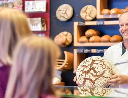 Ein Mann in einer Kochuniform hält ein rundes Stück Brot mit komplizierten Mustern. Hinter ihm ein Glasvitrine mit Brot und Regale mit mehr Brot. Zwei Frauen im Hintergrund schauen sich das Brot an.