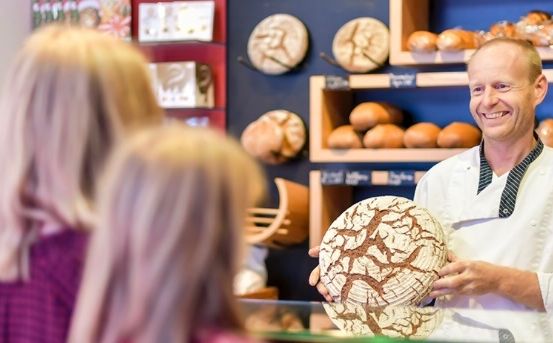 Ein Mann in einer Kochuniform hält ein rundes Stück Brot mit komplizierten Mustern. Hinter ihm ein Glasvitrine mit Brot und Regale mit mehr Brot. Zwei Frauen im Hintergrund schauen sich das Brot an.