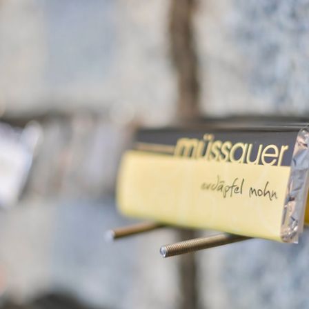 Eine gelbe Packung Müsli, beschriftet mit 'muessauer', ist an einem Metallhalter an einer Steinwand aufgehängt.