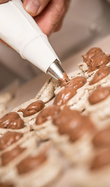 A hand uses a piping bag to apply chocolate onto cookies arranged on a wooden board.