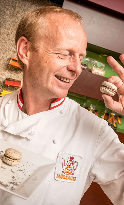 A chef wearing a white uniform smiles while holding a macaron. He is standing in front of a green shelf with several macarons.