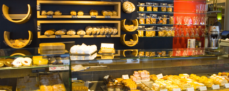 A bakery display case with various pastries, breads, and desserts on shelves. Glass shelves hold packaged snacks and glasses. A red pillar and a counter with more pastries are visible.