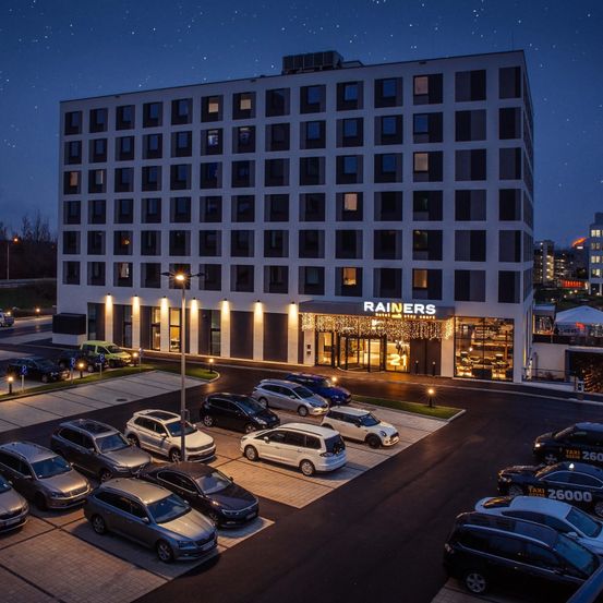 Das Rainers Hotel ist ein modernes Gebäude mit vielen Fenstern und Autos, die nachts davor geparkt sind. Der Eingang ist mit Lichtern beleuchtet und hat ein Schild darüber.