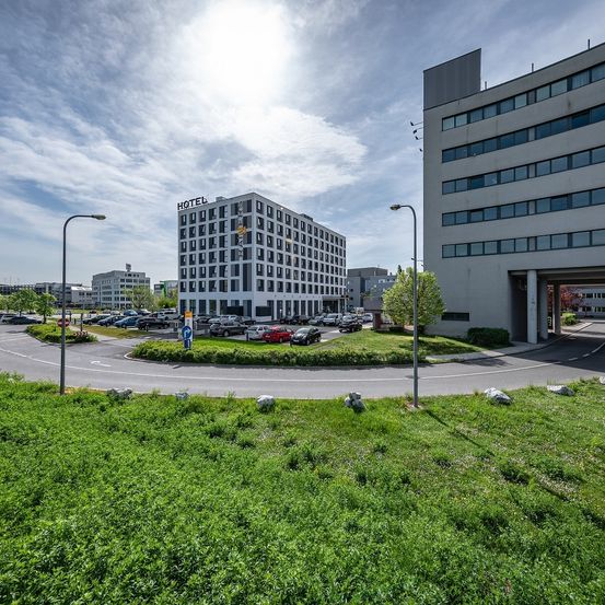 Ein modernes Hotel mit vielen Fenstern steht neben einem großen Gebäude. Mehrere Autos sind vor dem Hotel geparkt.