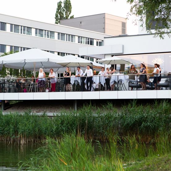 Eine Gruppe von Menschen steht unter Schirmen auf einem Balkon über einem Teich und blickt auf ein Gebäude.