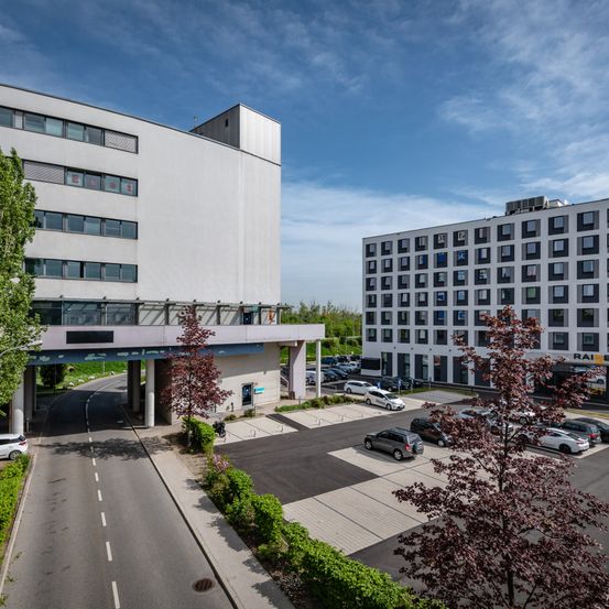 Eine Luftaufnahme eines modernen Gebäudekomplexes. Autos sind vor dem Gebäude mit vielen Fenstern geparkt, und andere stehen auf einem Parkplatz. Bäume und Büsche sind ebenfalls vorhanden.