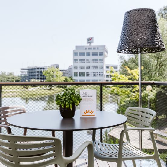 Ein Balkon mit einem runden Tisch und Stühlen, einem Topf mit Pflanzen und einer schwarzen Lampe. Dahinter ein See und ein Gebäude. Bäume und Pflanzen sind in der Umgebung.