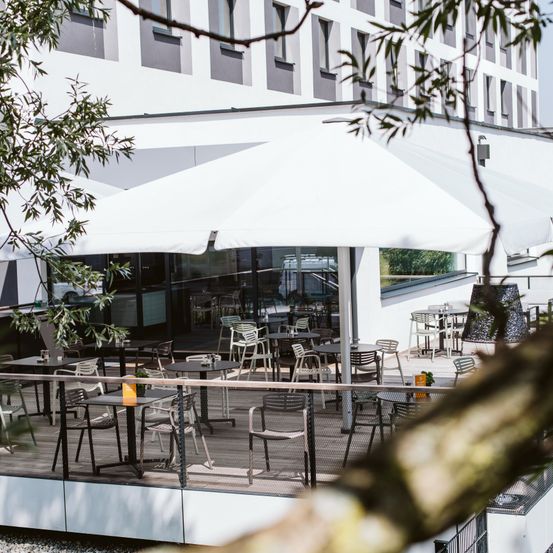 Eine Außenterrasse mit Tischen, Stühlen und einem großen weißen Sonnenschirm unter einem Gebäude mit Glasfenstern.