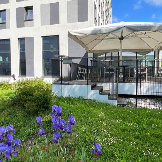 Eine Außenterrasse mit weißen Sonnenschirmen und Stühlen auf einem Balkon. Ein Rasenbereich mit lila Schwertlilien und weißen Blumen. Hinter dem Balkon befindet sich ein modernes Gebäude mit Glasfenstern.