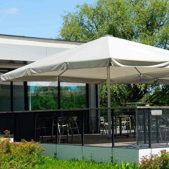 Ein großer weißer Sonnenschirm bedeckt eine Terrasse mit Tischen und Stühlen, umgeben von Pflanzen und einem Gebäude mit Glaswänden.