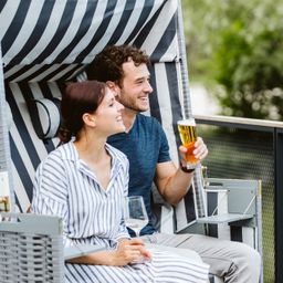 Ein Mann und eine Frau sitzen zusammen in einem schattigen Außenbereich, lächeln und halten Getränke. Die Frau trägt ein gestreiftes Kleid und der Mann ein blaues Hemd. Sie sitzen auf gepolsterten Stühlen mit einem Tisch in der Nähe.