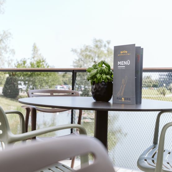 Ein runder Tisch mit Stühlen und einer Pflanze in einem schwarzen Topf. Ein Menü-Karte mit dem Wort 'Menü' darauf ist auf dem Tisch. Der Tisch ist auf einem Balkon mit einem Zaun und Blick auf ein Feld und Bäume.