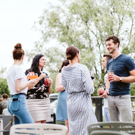 Eine Gruppe von Freunden, darunter Frauen und Männer, genießt Getränke und unterhält sich bei einem Outdoor-Treffen, umgeben von Bäumen und Tischen.