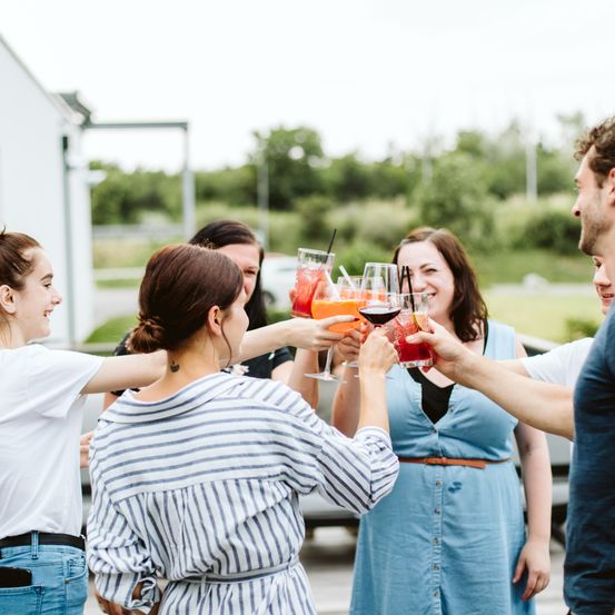 Mehrere Frauen und Männer sind draußen versammelt, lächeln und halten Weingläser. Sie scheinen ein Ereignis zu feiern, möglicherweise eine Hochzeit oder einen Geburtstag.
