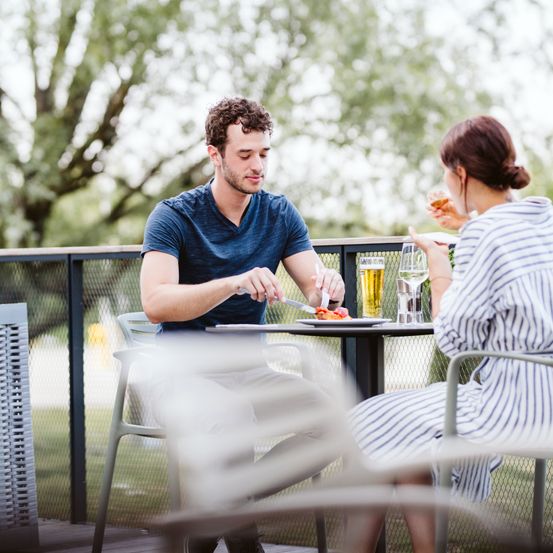 Ein Paar isst an einem Tisch im Freien, wobei der Mann seine Mahlzeit schneidet und die Frau einen Teller und ein Glas in der Hand hält. Sie sind von einem Zaun umgeben, mit Bäumen im Hintergrund.