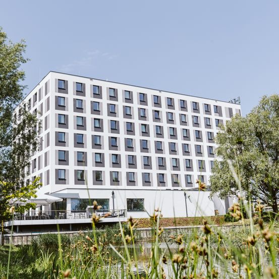 Ein modernes weißes und graues Gebäude mit vielen Fenstern und einem Balkon, umgeben von Bäumen und Gras.