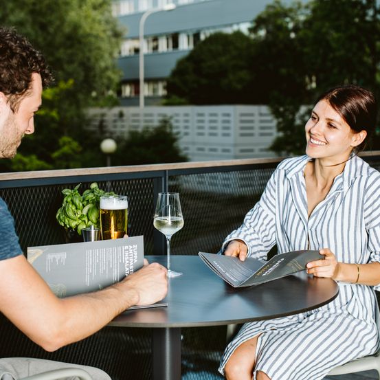 Ein Mann und eine Frau sitzen auf einem Balkon, lächeln und lesen eine Speisekarte. Sie haben Getränke und einen Topf auf ihrem Tisch.
