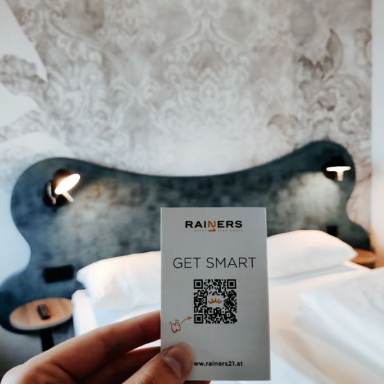 Eine Hand hält eine Karte mit 'RAINERS GET SMART' davor auf einem Bett. Die Tapete ist mit Mustern verziert.