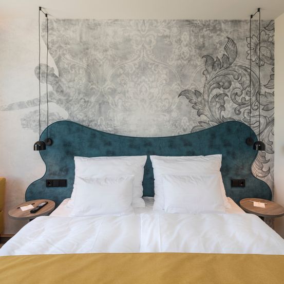 Ein ordentlich gemachtes Bett mit weißen Laken und blauem Kopfteil. Eine dekorative Wand dahinter mit verschnörkelten Mustern. Zwei Nachttische auf beiden Seiten mit Fernbedienungen und Karten.