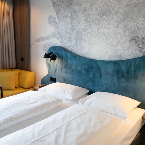 Ein Hotelzimmer mit zwei Einzelbetten, einem blauen Kopfteil und einer gemusterten Wand. Ein gelbes Sofa mit Kissen und Nachttischen ist ebenfalls vorhanden.