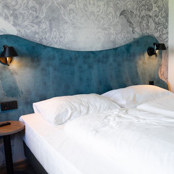 Ein ordentlich gemachtes Bett mit zwei weißen Kissen lehnt sich gegen ein blaues Kopfteil. Das Bett steht in der Nähe einer Wand mit dekorativer Tapete und zwei Wandlampen.