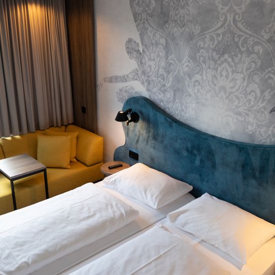 Ein gemütliches Hotelzimmer mit zwei Betten, einer gelben Couch und einer Wandmalerei. Das Zimmer hat ein blaues Kopfteil, weiße Kissen und dekorative Lampen.