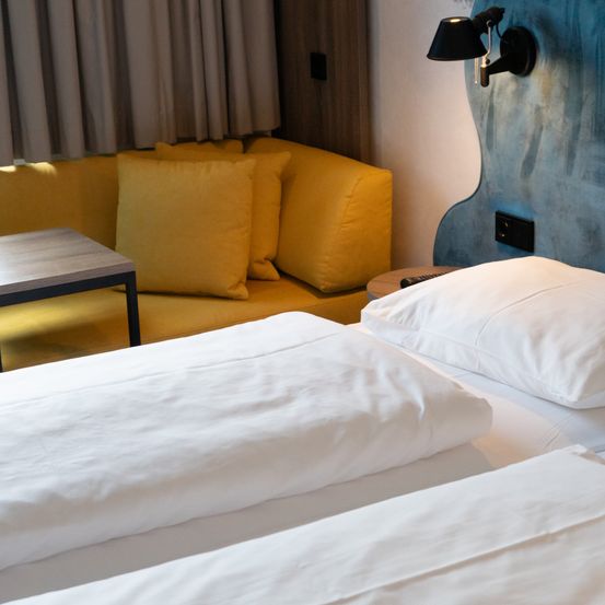 Ein Hotelzimmer mit einem Bett, einer gelben Couch und einem Kissen. Das Bett hat ein weißes Kissen und eine Lampe. Die Couch hat ein gelbes Kissen.