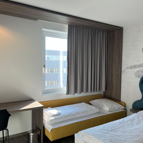 Ein einfaches Hotelzimmer mit zwei Einzelbetten, einem Schreibtisch und einem Fenster mit Vorhängen. Der Raum hat Holzwände und -böden.