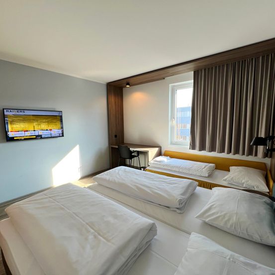 Ein sauberes und modernes Zimmer mit drei Betten, einem Fernseher, einem Schreibtisch und einem Fenster mit Vorhängen. Das Zimmer ist hell und gut beleuchtet.