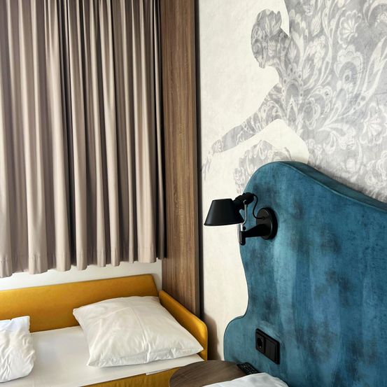 Ein Schlafzimmer mit einem in gelbem Stoff bezogenen Bett, zwei weißen Kissen, einem blauen Kopfteil mit Wandlampe und einer gemusterten Wand mit einer Silhouette.