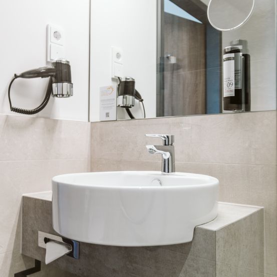 Ein Badezimmer mit einem modernen Waschbecken, Spiegel und an der Wand montiertem Föhn, zusammen mit einer Flasche Shampoo.