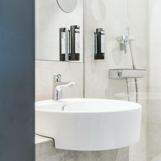 Ein modernes Badezimmer mit einem weißen Waschbecken, silbernem Wasserhahn und Glasdusche. Drei schwarze Seifenspender sind an der Wand montiert.