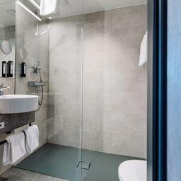 Ein Badezimmer mit grauen Fliesen und einer Glasdusche. Ein Waschbecken, ein Spiegel und ein Handtuchhalter sind sichtbar. Ein WC und Handtücher befinden sich rechts.