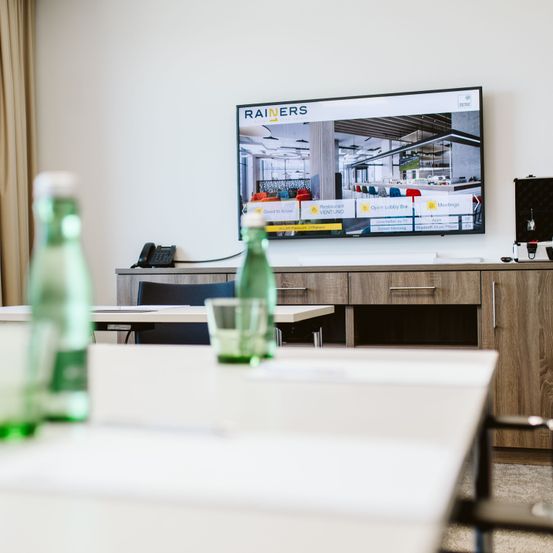 Ein Konferenzraum mit einem großen Bildschirm, der Rainers, einen Konferenzraum und ein Restaurant zeigt. Zwei leere Tische haben Flaschen und Gläser. Ein Holzschrank hält ein Telefon.