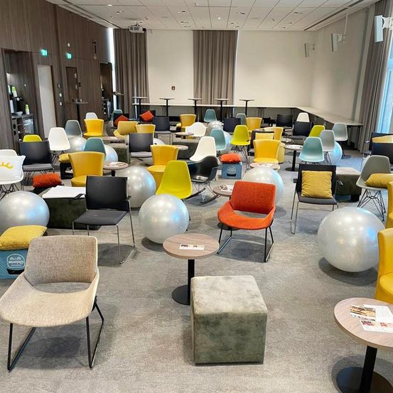 Ein Innenraum mit verschiedenfarbigen Stühlen, Tischen und Fitnessbällen. Der Raum hat einen Teppichboden und einen Projektor an der Decke.
