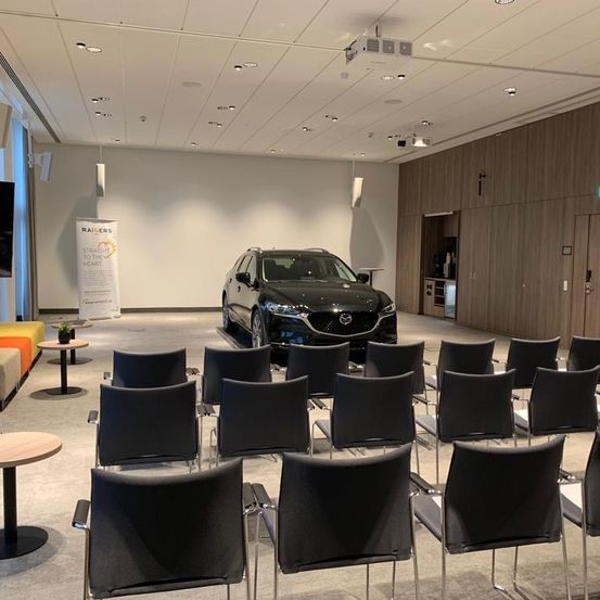 Ein leerer Konferenzraum mit Reihen schwarzer Stühle, einem Tisch, einem schwarzen Auto in der Mitte, einer Holzwand, einem Fernseher und einem hölzernen Schild.