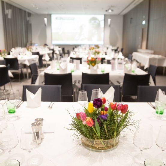 Ein Konferenzraum, der für eine Veranstaltung vorbereitet ist, mit Blumenarrangements auf Tischen und einer Projektionsleinwand vorne.