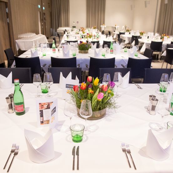 Ein gut organisierter Bankettsaal mit Tischen und Stühlen. Die Tische haben weiße Tischdecken, Blumenarrangements, Weingläser, Flaschen und Besteck. Der Raum hat Vorhänge und Lichter an der Decke.