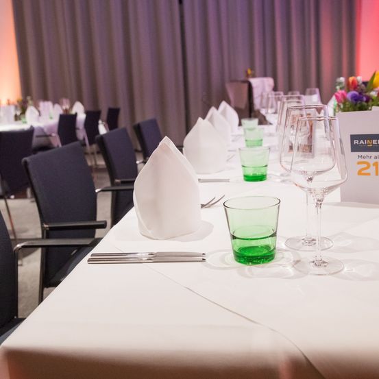 Ein langer Esstisch für eine formelle Veranstaltung. Jede Platzkarte beinhaltet einen grünen Becher, ein Weinglas und Besteck. Der Tisch ist mit einer weißen Tischdecke bedeckt.