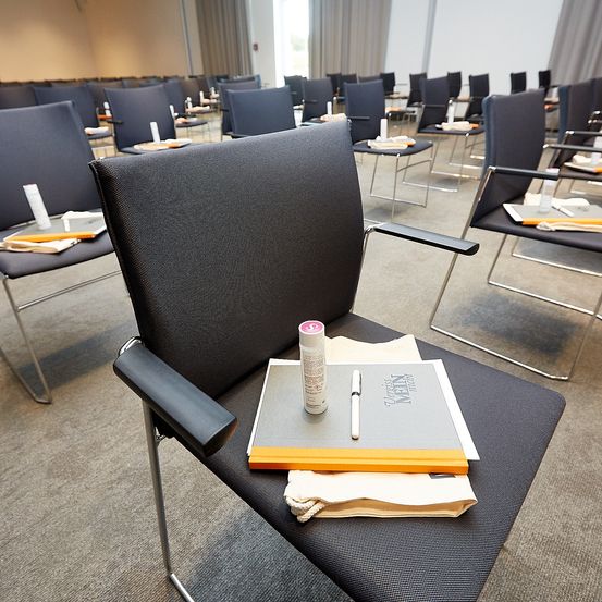 Ein Konferenzraum mit Reihen von Stühlen. Ein schwarzer Stuhl hat ein Buch, einen Stift und eine Flasche darauf. Alle Stühle sind ordentlich aufgestellt mit einer Kerze und einem Teller auf jedem.