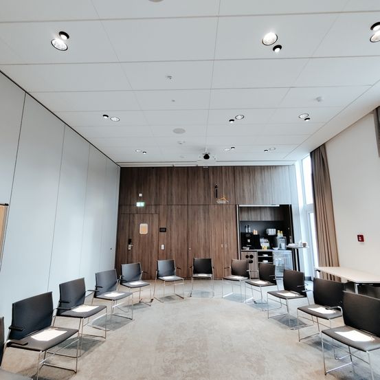 Ein leerer Besprechungsraum mit Stühlen im Kreis angeordnet, Holzwänden und einer weißen Decke mit Einbauleuchten.